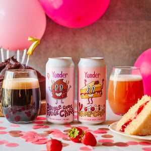 Two cans of Yonder Double Chocolate Fudge Cake beer with a slice of cake, glass of dark beer, and glass of light beer on a table with pink balloons and cake in the background.