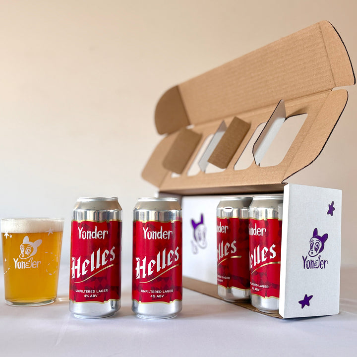 Yonder Helles beer cans and a glass on a white surface with a cardboard carrier.