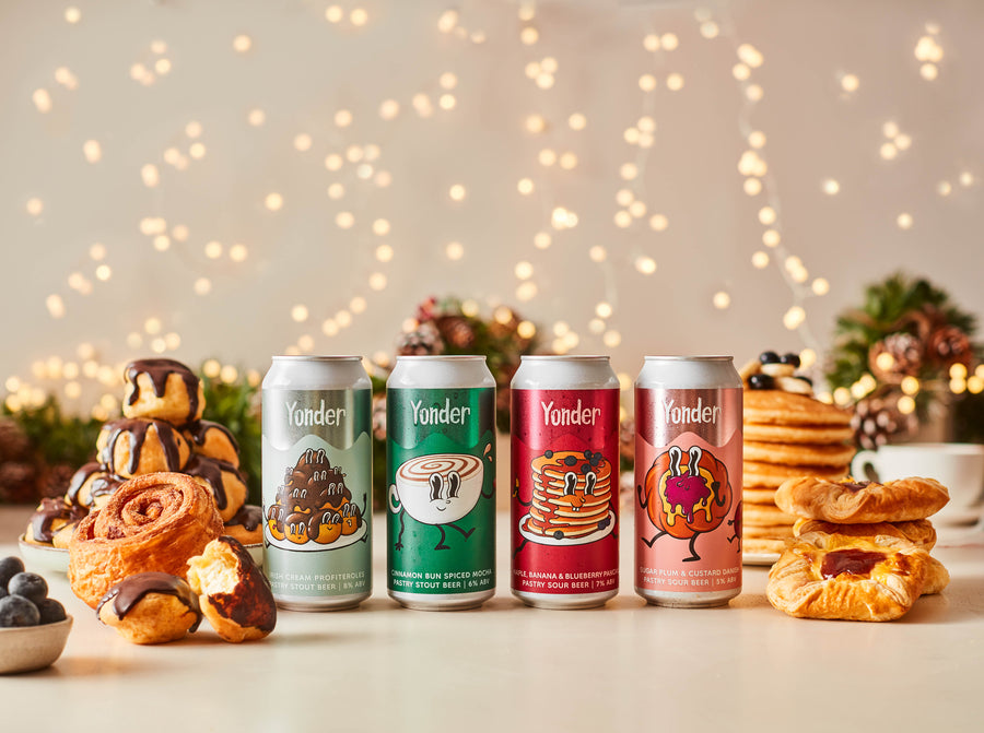 Four cans of Yonder beer with colorful labels on a table with pastries and blurred lights in the background