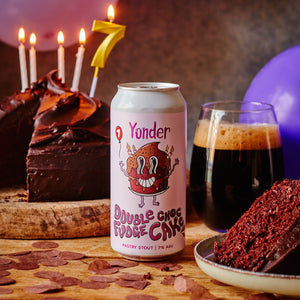 Yonder Double Choc Fudge Cake beer can with a chocolate cake, glass of dark beer, and candles on a wooden table.